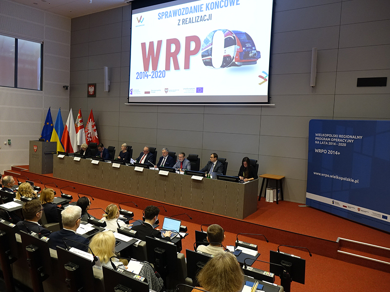 Zdjęcie wykonano w sali audytoryjnej. Na scenie, za długim pulpitem siedzi osiem osób. Nad nimi znajduje się ekran z napisem: Sprawozdanie końcowe z realizacji WRPO 2014-2020. Wśród publiczności znajduje się kilkadziesiąt osób. Każdy ma do dyspozycji własny pulpit z mikrofonem. Fotografia pochodzi z archiwum Urzędu Marszałkowskiego Województwa Wielkopolskiego.