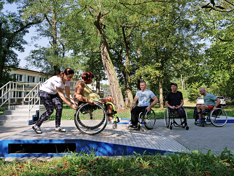 Na zdjęciu znajdują się cztery osoby na wózkach inwalidzkich, które przy wsparciu asystenta trenują pokonywanie przeszkód w specjalnie do tego zaprojektowanej przestrzeni w parku. Fotografia pochodzi z archiwum beneficjenta.
