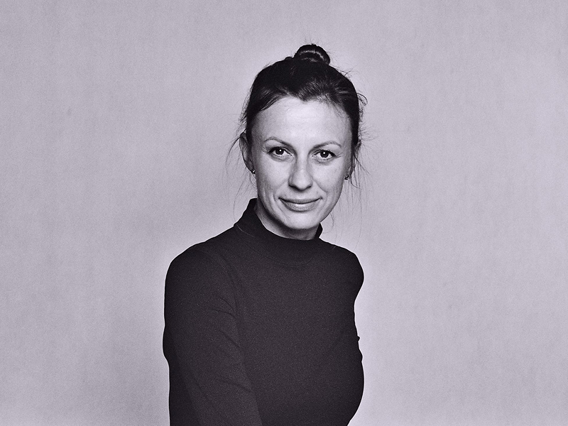 Czarno-biała fotografia przedstawia uśmiechniętą kobietę w średnim wieku, o upiętych, ciemnych włosach, w swetrze. 