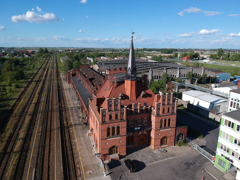 Fotografia z lotu ptaka przedstawia dworzec kolejowy, przypominający nieco w swojej budowie pałac – ma strzelistą wieżę, jest z czerwonej cegły. Z lewej znajdują się tory kolejowe, po prawej zaś zabudowania miejskie. Autorem zdjęcia jest Aleksander Liebert.