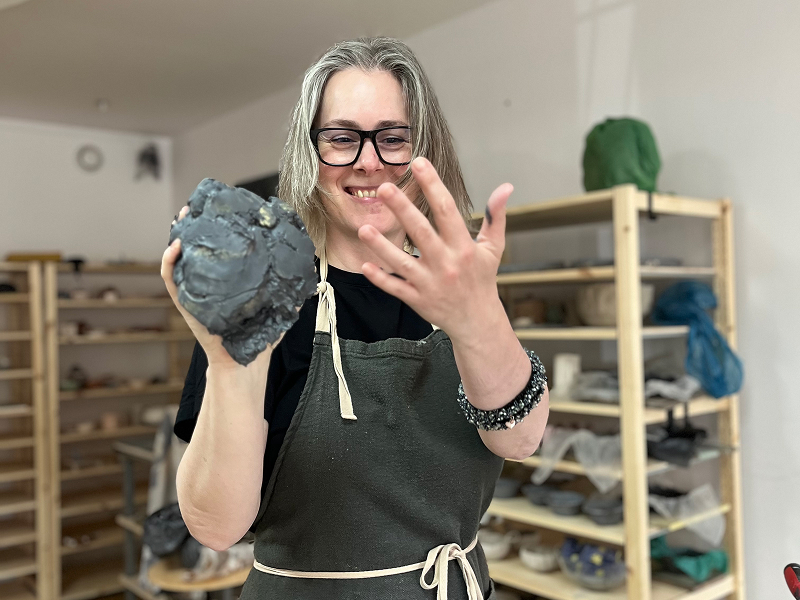 Fotografia przedstawia uśmiechniętą kobietę o jasnych włosach, która urabia glinę. Na sobie ma szary fartuch. Zdjęcie pochodzi z archiwum Ubabrane.