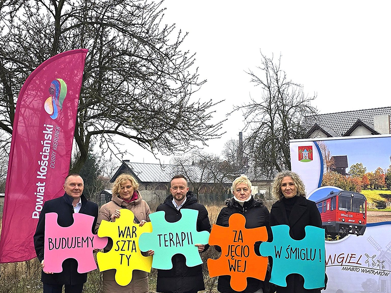 Na ilustracji widocznych jest pięć dorosłych osób, stojących obok siebie, na świeżym powietrzu. Każda z nich trzyma jeden kolorowy puzzel, z wyrazem. Napis układa się w całość: Budujemy warsztaty terapii zajęciowej w Śmiglu.
