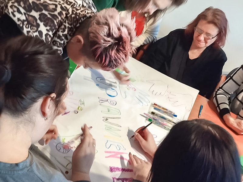 Scena przedstawia warsztaty dla kobiet. Panie siedzą przy stoliku i tworzą plakat z napisem „równouprawnienie”. Fotografia pochodzi z archiwum beneficjenta.