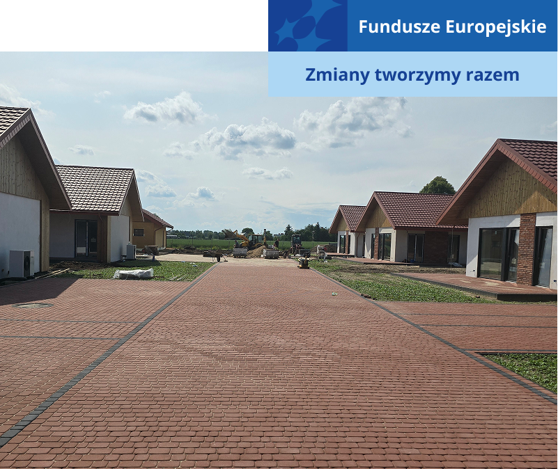 Ilustracja ukazuje brukowaną drogę, przy której, po obu stronach, stoją parterowe domki w trakcie budowy. Zdjęcie pochodzi z archiwum beneficjenta.