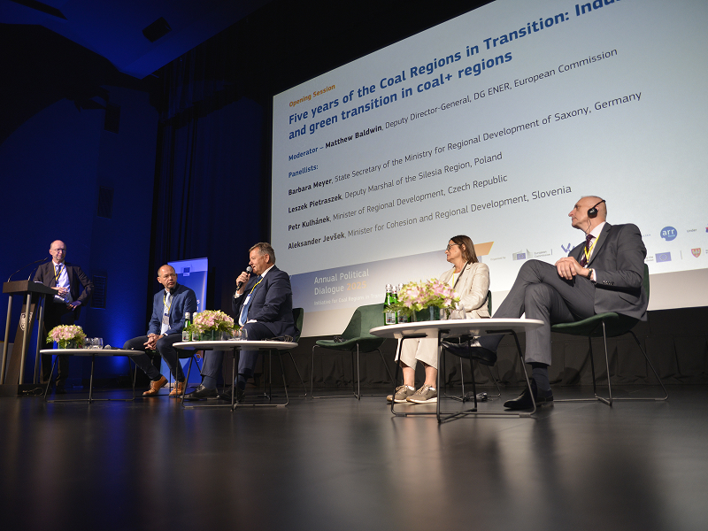Zdjęcie przedstawia debatę w trakcie konferencji. Na scenie siedzi czterech ekspertów – trzech mężczyzn i kobieta. Jeden z mężczyzn przemawia do mikrofonu. Z boku stoi prowadzący. Autorem fotografii jest Dominik Wójcik.
