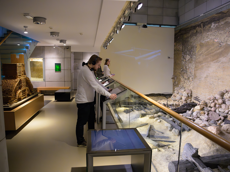 Fotografię wykonano w jednej z sal muzeum archeologicznego. Po prawe, za barierkami, widoczne są odkryte wykopaliska. Z lewej znajduje się zaś drewniany schemat. Widoczni są również zwiedzający. Zdjęcie pochodzi z archiwum beneficjenta.
