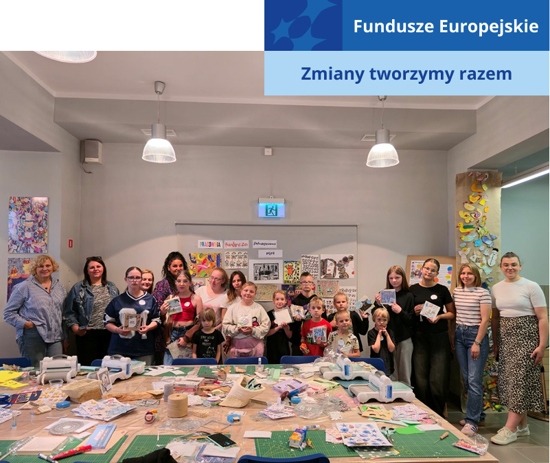 Zdjęcie przedstawia sporą grupę osób, w różnym wieku, kobiet i mężczyzn, którzy pozują po zakończonych warsztatach. Niektórzy prezentują efekty swojej pracy. Przed nimi znajduje się duży stół z materiałami i narzędziami. Fotografia pochodzi z archiwum beneficjenta.