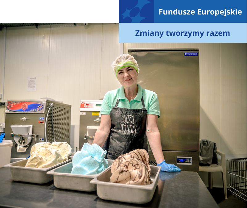 Na zdjęciu znajduje się uśmiechnięta kobieta w ochronnym czepku, fartuchu i rękawicach. Przed nią, na stalowym stole, stoją trzy pudła z kolorowymi lodami. Autorem fotografii jest Dominik Wójcik.