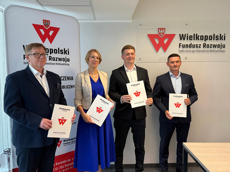 Ilustracja prezentuje cztery osoby – trzech mężczyzn i kobietę, którzy prezentują podpisane umowy. Pozują na tle ścianki z napisem „Wielkopolski Fundusz Rozwoju”. Na dole zdjęcia znajduje się belka z logotypami: Funduszy Europejskich dla Wielkopolski, Rzeczpospolitej Polskiej, Dofinansowane przez Unię Europejską oraz, oddzielone pionową kreską, Samorządu Województwa Wielkopolskiego. Fotografia pochodzi z archiwum Wielkopolskiego Funduszu Rozwoju.