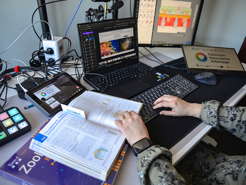 Fotografia ukazuje stanowisko do zdalnego prowadzenia lekcji. Widoczny jest monitor, laptop, klawiatura, myszka, kamery i mikrofon. Na biurku znajdują się również podręczniki. Autorem zdjęcia jest Dominik Wójcik.