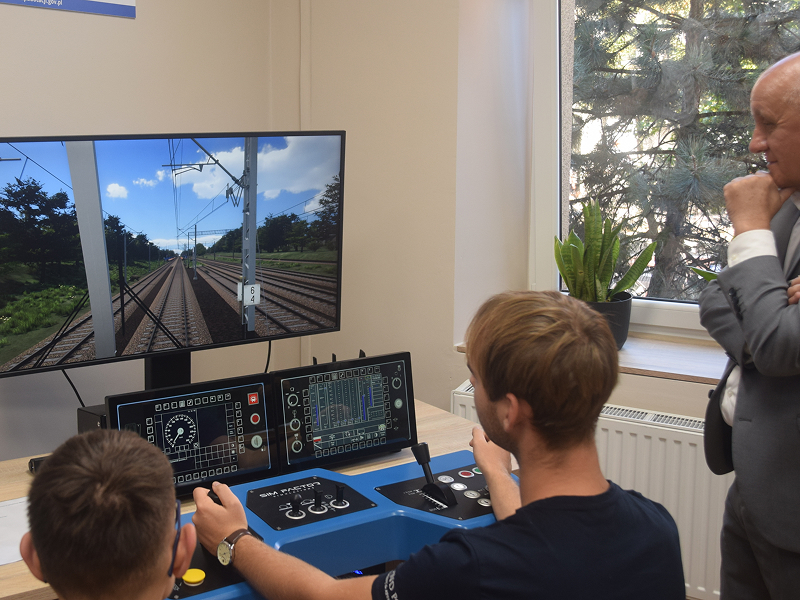 Na zdjęciu widocznych jest dwóch uczniów, którzy pod nadzorem stojącego nad nimi nauczyciela, obsługują symulator kolejowy. Urządzenie składa się z monitora przedstawiającego widok z wnętrza kabiny lokomotywy oraz panelu do sterowania. Fotografia pochodzi z archiwum beneficjenta.