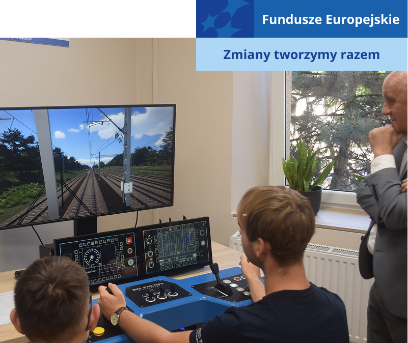 Na zdjęciu widocznych jest dwóch uczniów, którzy pod nadzorem stojącego nad nimi nauczyciela, obsługują symulator kolejowy. Urządzenie składa się z monitora przedstawiającego widok z wnętrza kabiny lokomotywy oraz panelu do sterowania. Fotografia pochodzi z archiwum beneficjenta.