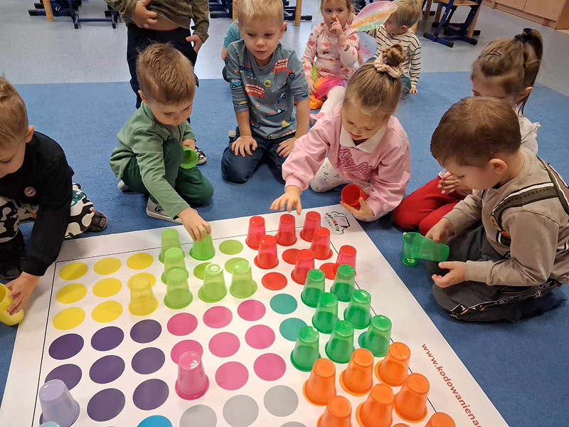 Zdjęcie ukazuje grupę przedszkolaków, którzy klęczą przed planszą rozłożoną na dywanie. Na planszy znajdują się liczne kolorowe pola. Dzieci ustawiają na nich kolorowe kubki. Fotografia pochodzi z archiwum beneficjenta.