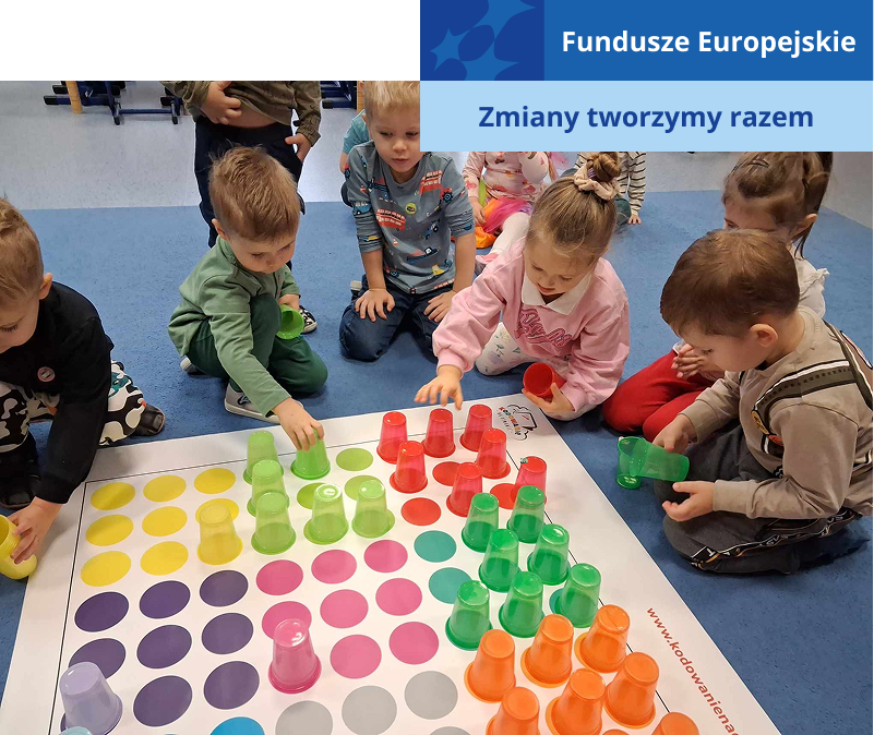 Zdjęcie ukazuje grupę przedszkolaków, którzy klęczą przed planszą rozłożoną na dywanie. Na planszy znajdują się liczne kolorowe pola. Dzieci ustawiają na nich kolorowe kubki. Fotografia pochodzi z archiwum beneficjenta.