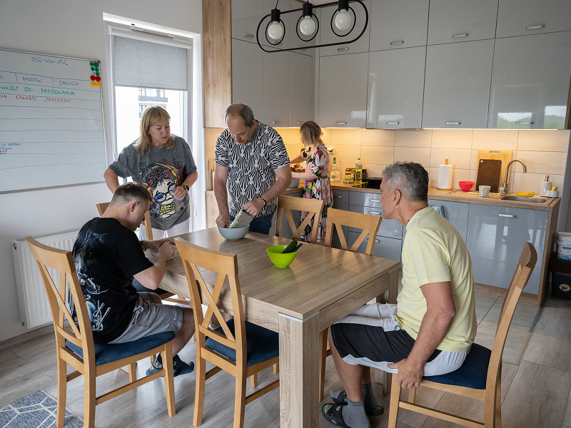 Fotografia przedstawia sytuację w mieszkaniu wspomaganym. Widać na nim dwie kobiety i trzech mężczyzn w kuchni. Panie pomagają panom przygotować posiłek. Autorem zdjęcia jest Maciej Motylewski.