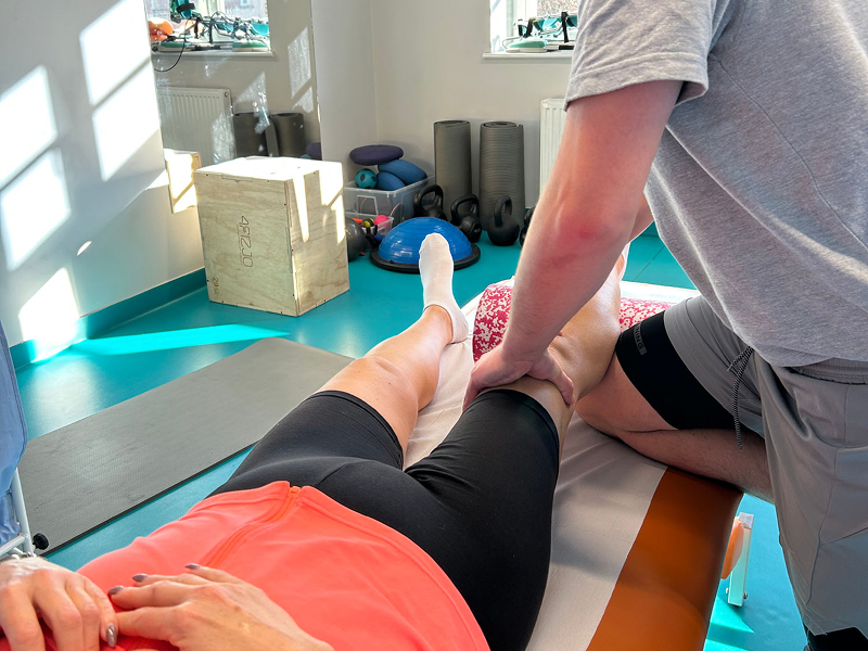 Fotografia przedstawia fizjoterapeutę, który masuje prawą łydkę pacjentki, leżącej na kozetce. Zdjęcie pochodzi z archiwum beneficjenta.