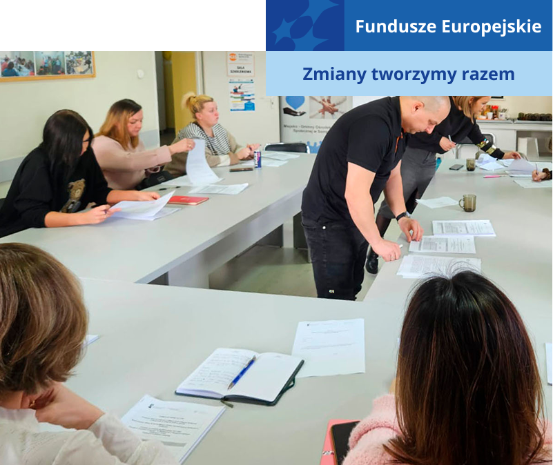 Zdjęcie wykonano w trakcie szkolenia. Przy stoliku w kształcie podkowy siedzi kilka kobiet. W środku krąży dwóch prowadzących – kobieta i mężczyzna. W tyle widać biurko i tablicę. Fotografia pochodzi z archiwum beneficjenta.