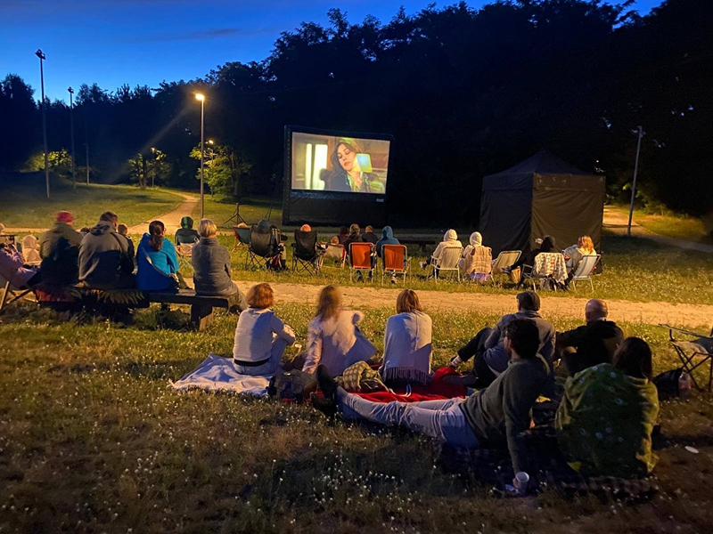 Zdjęcie wykonano późnym wieczorem w parku. W tle widoczny jest ekran, na którym wyświetlany jest film. Wokół, na kocach siedzą ludzie. Fotografia pochodzi z archiwum beneficjenta.