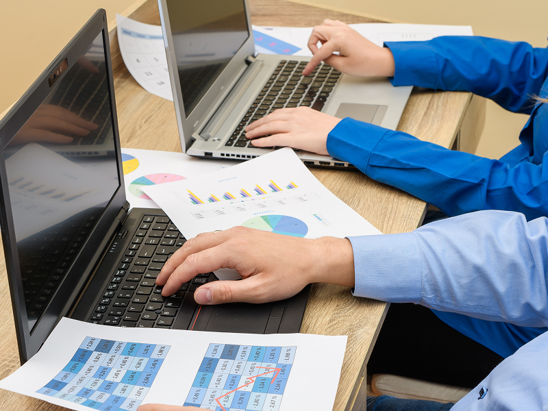 Fotografia przedstawia dwa laptopy, które obsługiwane są przez dwie osoby. Na ekranach widać skomplikowane wykresy. Wokół leżą również wydruki analiz. Zdjęcie pochodzi z adobe stock.