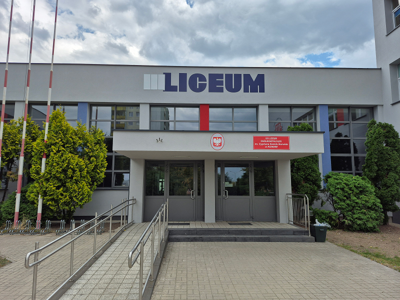 Zdjęcie przedstawia wejście do szkoły. Widoczne są podwójne drzwi, do których prowadzą schody oraz podjazd dla osób z niepełnosprawnością. Elewacja jest szara. Na krawędzi budynku widoczny jest napis Trzecie Liceum. Fotografia pochodzi z archiwum beneficjenta.