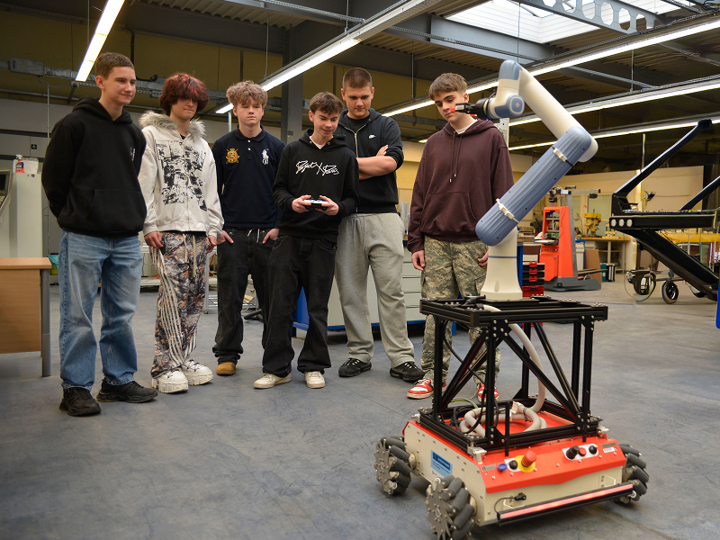 Zdjęcie wykonano w laboratorium maszyn inteligentnych. Sześciu chłopaków przygląda się zdalnie sterowanemu robotowi. Jeden z nich trzyma kontroler. Z prawej strony widoczny jest plakat z napisem Wielkopolska, na niebieskim tle. Autorem fotografii jest Dominik Wójcik.