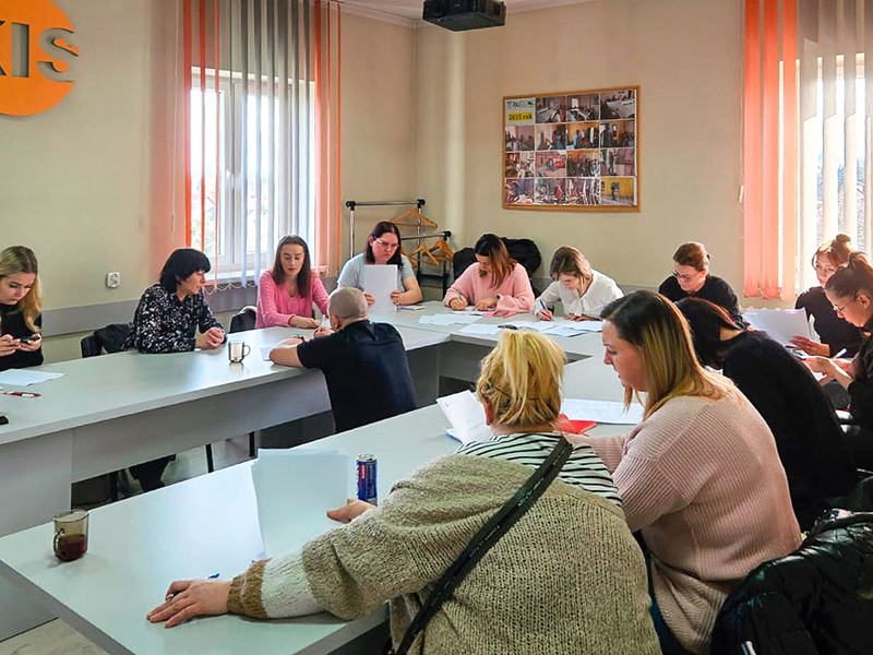 Zdjęcie wykonano w trakcie kursu dla kobiet. Panie siedzą przy stole w kształcie podkowy. W środku znajduje się mężczyzna – prowadzący zajęcia. Fotografia pochodzi z archiwum beneficjenta.
