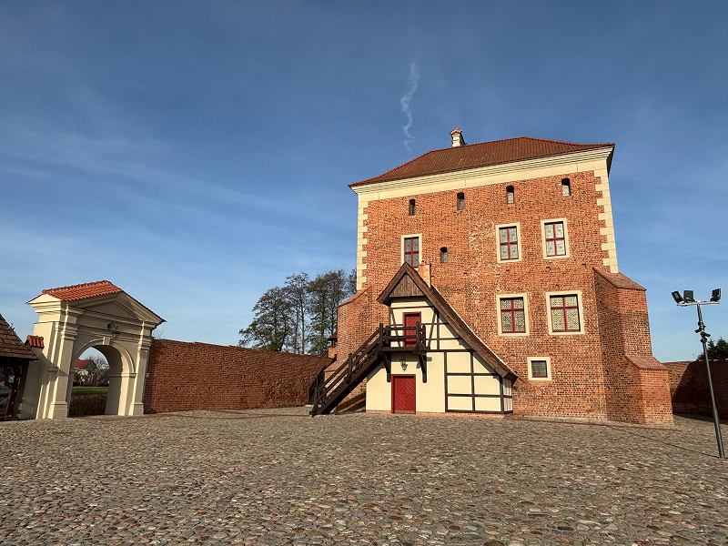 Na zdjęciu widoczne jest jezioro. Na jego dalekim brzegu widoczna jest wieża i kilka zamkowych zabudowań (autorem zdjęcia jest Sławomir Maciaszek)