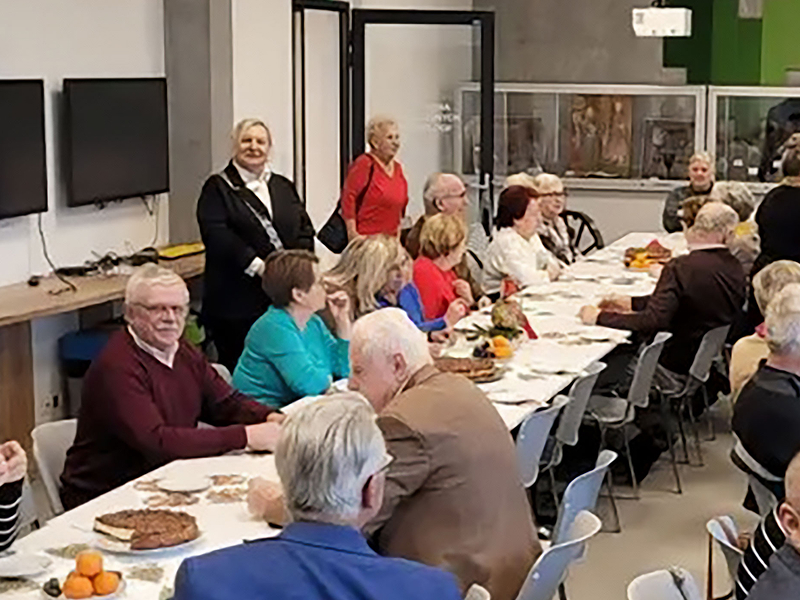 Na ilustracji widać sporą grupę seniorów siedzących przy dwóch długich stołach, na których znajdują się między innymi desery. Zdjęcie pochodzi z archiwum beneficjenta.