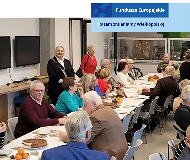 Na ilustracji widać sporą grupę seniorów siedzących przy dwóch długich stołach, na których znajdują się między innymi desery. Zdjęcie pochodzi z archiwum beneficjenta.