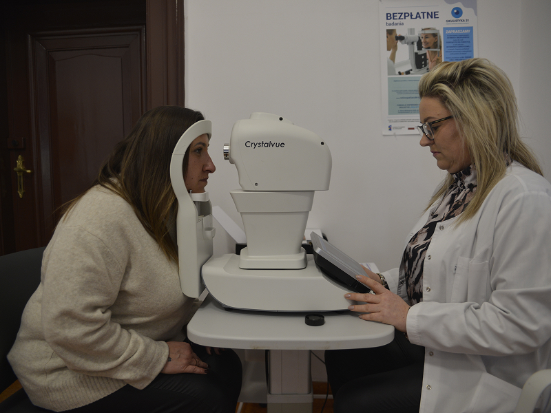 Zdjęcie wykonano w gabinecie lekarski. Na przeciwko siebie siedzą dwie kobiety. Po prawej osoba wykonująca badanie, w białym fartuchu, wpisuje komendy na kontrolerze. Po lewej pacjentka, z głową umieszczoną w czytniku urządzenia. Sprzęt znajduje się pomiędzy nimi. Autorem fotografii jest Dominik Wójcik.