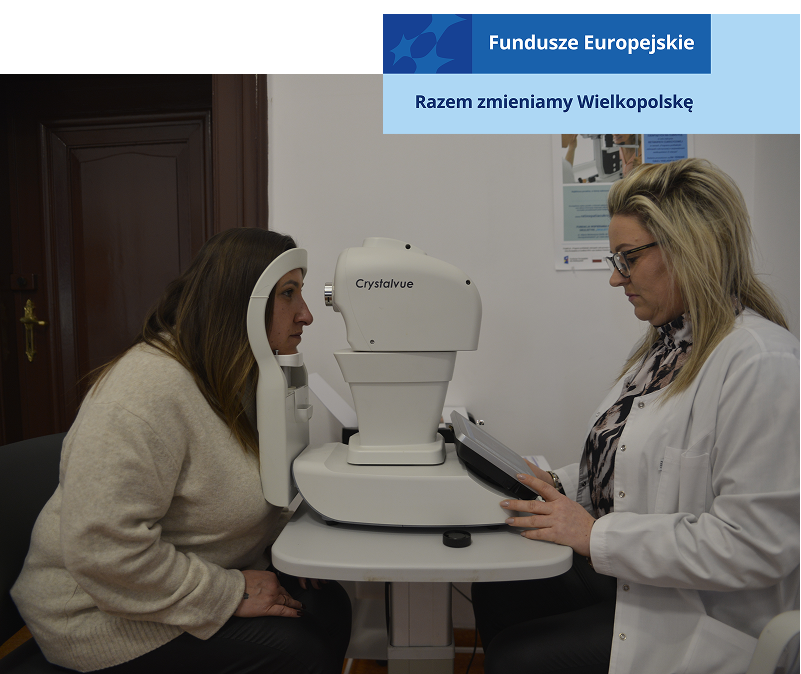 Zdjęcie wykonano w gabinecie lekarski. Na przeciwko siebie siedzą dwie kobiety. Po prawej osoba wykonująca badanie, w białym fartuchu, wpisuje komendy na kontrolerze. Po lewej pacjentka, z głową umieszczoną w czytniku urządzenia. Sprzęt znajduje się pomiędzy nimi. Autorem fotografii jest Dominik Wójcik.