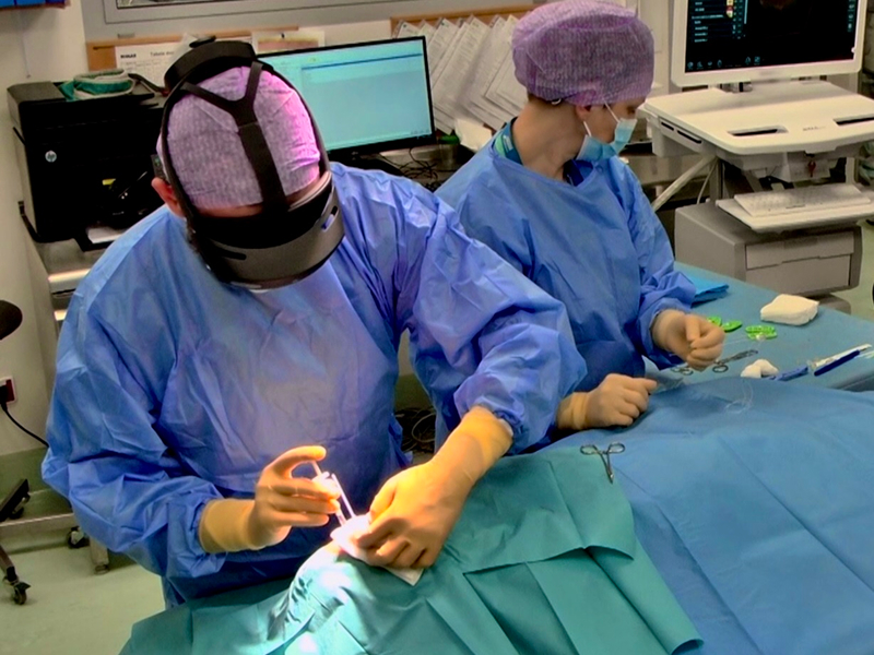 Zdjęcie przedstawia chirurgów w trakcie operacji. Ubrani są w ochronne stroje i czepki. Jeden z nich ma założone google wirtualnej rzeczywistości. Fotografia pochodzi z archiwum beneficjenta.