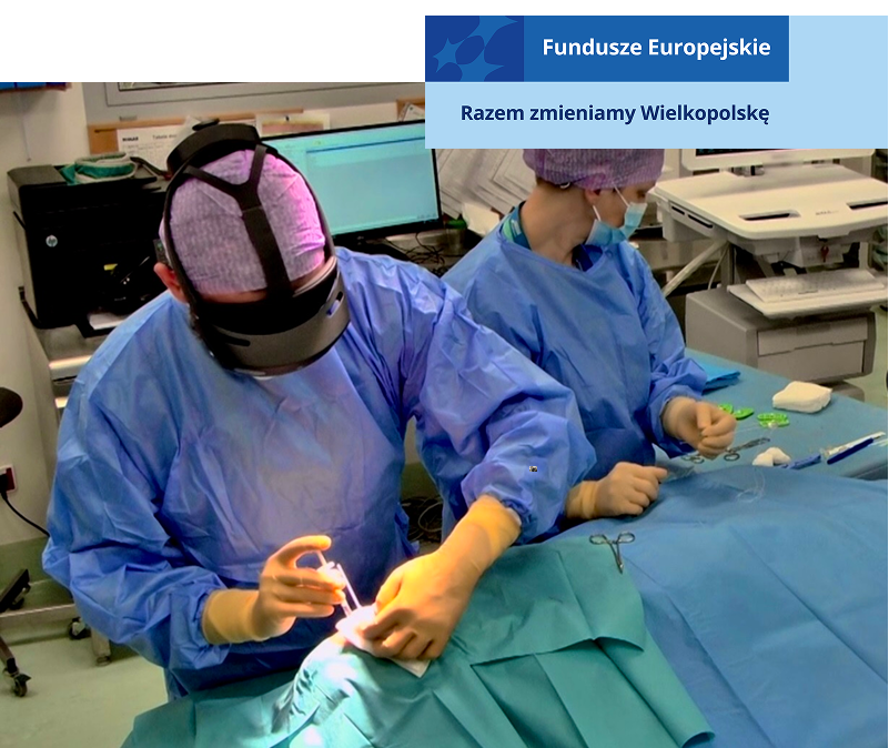 Zdjęcie przedstawia chirurgów w trakcie operacji. Ubrani są w ochronne stroje i czepki. Jeden z nich ma założone google wirtualnej rzeczywistości. Fotografia pochodzi z archiwum beneficjenta.