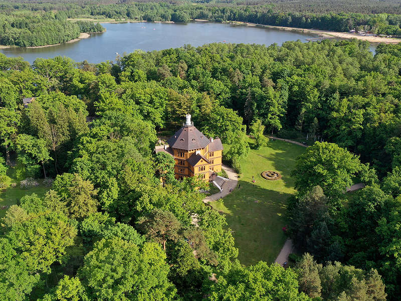 Zdjęcie z lotu ptaka przedstawia położony w środku lasu dworek, za którym znajduje się sporych rozmiarów jezioro. Fotografia pochodzi z archiwum beneficjenta.