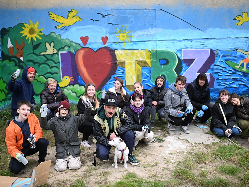Fotografia ukazuje sporą grupę młodych ludzi i pieska, którzy z wyraźną radością i ekscytacją pozują przed ścianą przyozdobioną muralem. Osoby znajdujące się na zdjęciu trzymają puszki z farbą, więc to one są autorami muralu. Sam malunek przedstawia jezioro i jego brzeg. Widoczna jest osoba pływająca na desce oraz kilka zwierząt. Na środku widoczny jest napis: I, symbol serca, Trz. Zdjęcie pochodzi z archiwum beneficjenta.