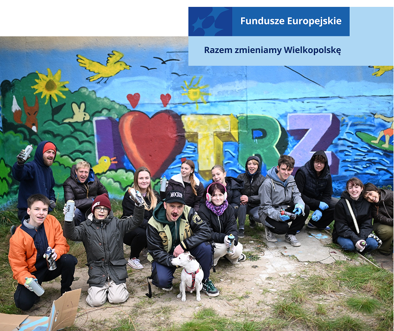 Fotografia ukazuje sporą grupę młodych ludzi i pieska, którzy z wyraźną radością i ekscytacją pozują przed ścianą przyozdobioną muralem. Osoby znajdujące się na zdjęciu trzymają puszki z farbą, więc to one są autorami muralu. Sam malunek przedstawia jezioro i jego brzeg. Widoczna jest osoba pływająca na desce oraz kilka zwierząt. Na środku widoczny jest napis: I, symbol serca, Trz. Zdjęcie pochodzi z archiwum beneficjenta.