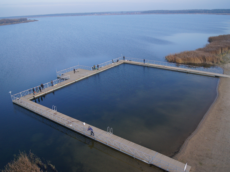 Na zdjęciu znajduje się kwadratowy pomost na jeziorze, po którym chodzą ludzie. Jest słoneczna pogoda. Fotografia pochodzi z archiwum beneficjenta.