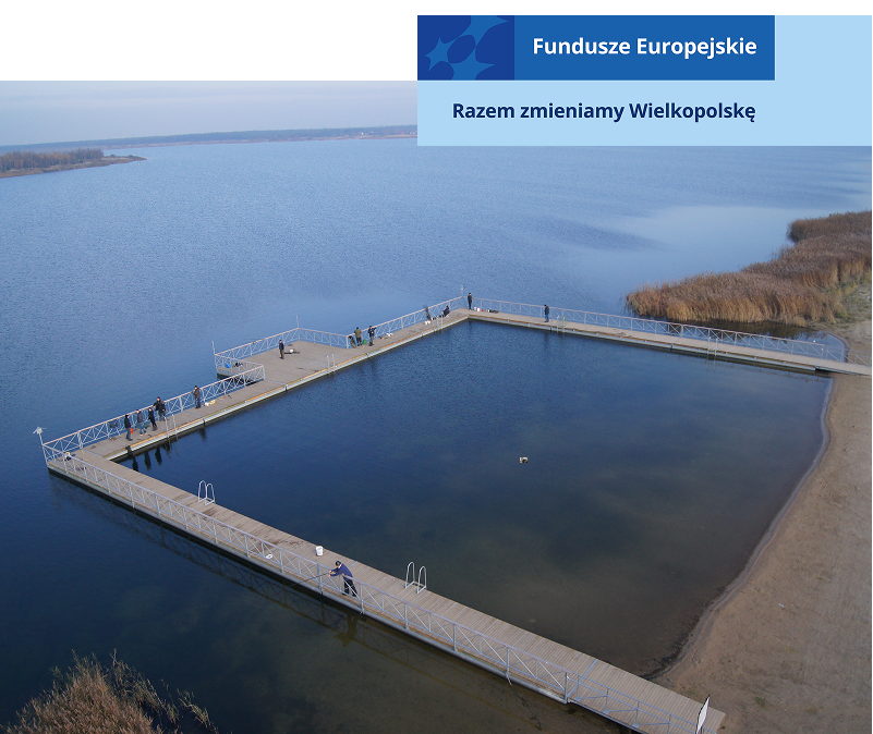 Na zdjęciu znajduje się kwadratowy pomost na jeziorze, po którym chodzą ludzie. Jest słoneczna pogoda. Fotografia pochodzi z archiwum beneficjenta.