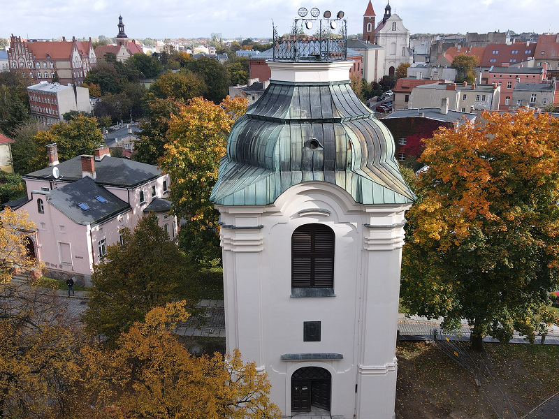 Zdjęcie z lotu ptaka przedstawia zbliżenie na kamienną wieżę, która jest dzwonnicą. W tle widoczne są miejskie zabudowania. Fotografia pochodzi z archiwum beneficjenta.