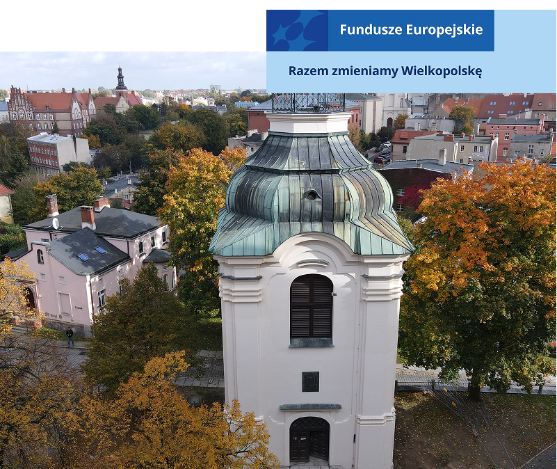 Zdjęcie z lotu ptaka przedstawia zbliżenie na kamienną wieżę, która jest dzwonnicą. W tle widoczne są miejskie zabudowania. Fotografia pochodzi z archiwum beneficjenta.