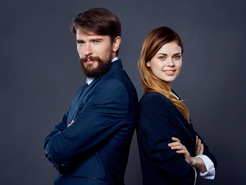 Zdjęcie przedstawia dwie elegancko ubrane osoby – kobietę i mężczyznę, stojących do siebie plecami, na ciemnym tle. Fotografia pochodzi z adobe stock.