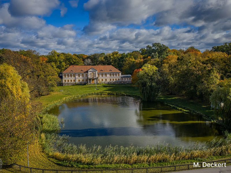 Zdjęcie przedstawia pałac, stojący wokół drzew. Przed budynkiem znajduje się staw. Autorem fotografii jest M. Deckert.