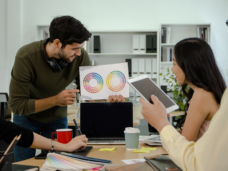 Zdjęcie przedstawia czwórkę młodych osób – dwie kobiety i dwóch mężczyzn pracujących nad projektem w firmie. Jeden z mężczyzn trzyma schemat, pozostali przyglądają się. Widoczne są rzeczy na stole: laptop, przybory do pisania. Fotografia pochodzi z adobe stock.