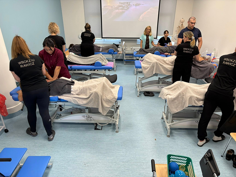 Fotografia przedstawia kurs masażysty, prowadzony w nowoczesnej sali. Widoczni są pacjenci leżący na kozetkach, instruktorzy oraz kursanci. Zdjęcie pochodzi z archiwum beneficjenta.