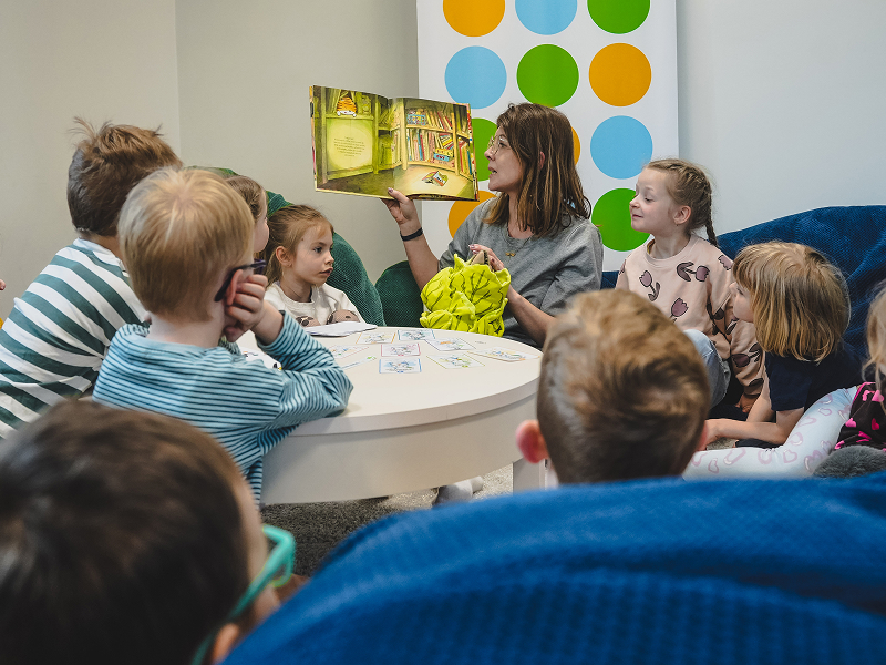 Zdjęcie przedstawia grupę dzieci, które z uwagą słuchają bajki czytanej przez opiekunkę. Autorką fotografii jest Zuzanna Kaczmarek z Urzędu Gminy Dopiewo.