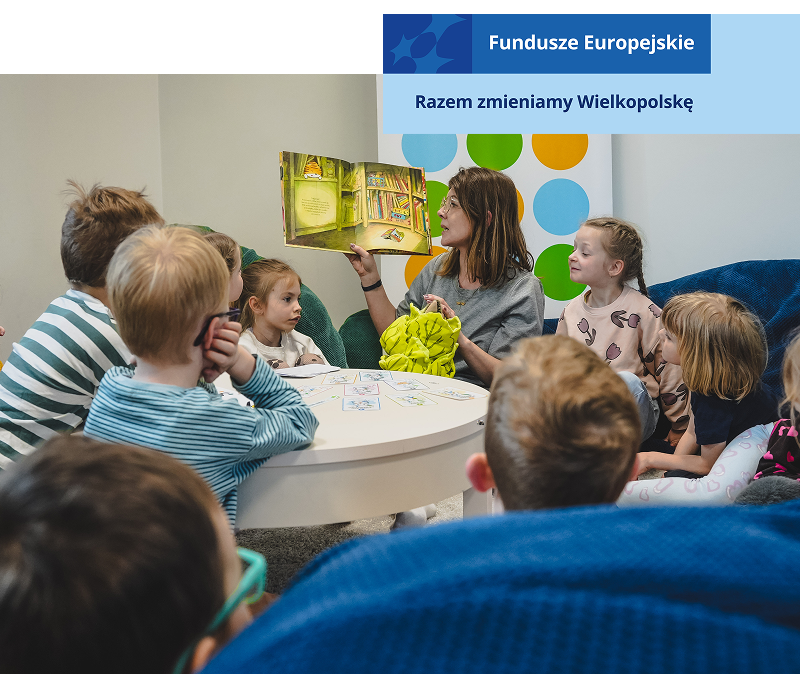 Zdjęcie przedstawia grupę dzieci, które z uwagą słuchają bajki czytanej przez opiekunkę. Autorką fotografii jest Zuzanna Kaczmarek z Urzędu Gminy Dopiewo.