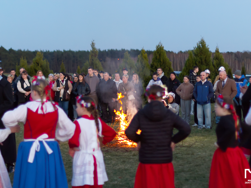 Fotografię wykonano na łące. Spora grupa ludzi, z których część jest w tradycyjnych, ludowych strojach, zgromadziła się wokół ogniska. Zdjęcie pochodzi z archiwum Muzeum Ziemi Rawickiej.