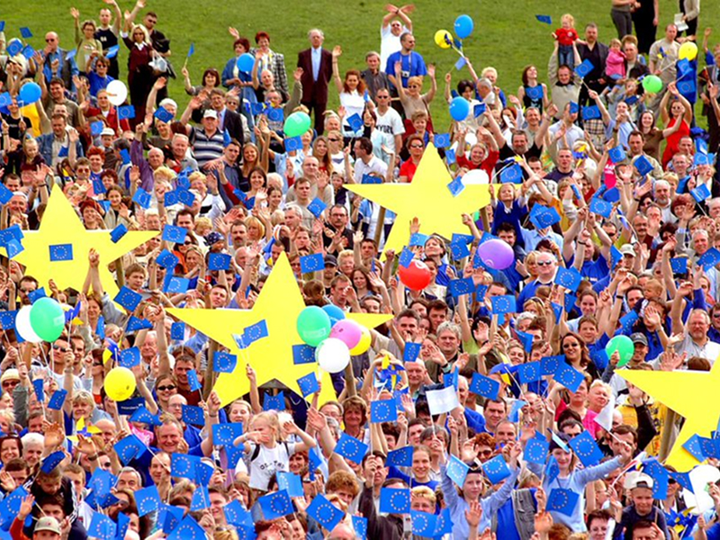 Na zdjęciu widoczny jest tłum ludzi stojących na trawie trzymających unijne flagi, balony czy żółte gwiazdy. Fotografia pochodzi z archiwum Urzędu Marszałkowskiego Województwa Wielkopolskiego.