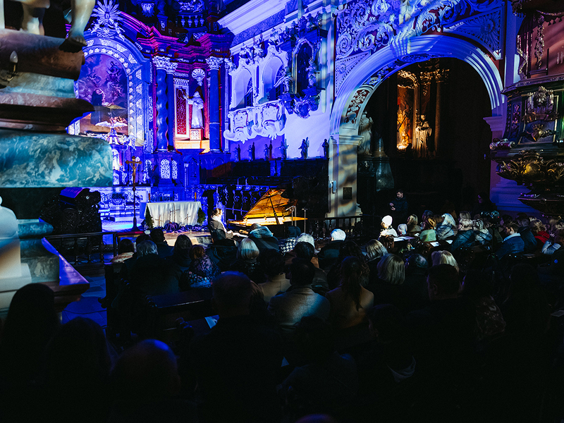 Zdjęcie przedstawia koncert fortepianowy odbywający się w kościele. Widoczne są imponujące zdobienia, figury. Solista zasiada przy fortepianie ustawionym przed ołtarzem. Publiczność znajduje się w dalszej części wnętrza. Pomieszczenie skąpane jest w ciemnych i niebieskich barwach. Fotografia pochodzi z archiwum beneficjenta.