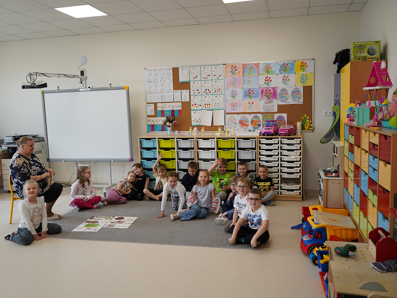 Fotografia przedstawia grupkę przedszkolaków siedzących na dywanie w sali. Obok, na krześle, siedzi nauczycielka. Autorem zdjęcia jest Sławomir Dawid.